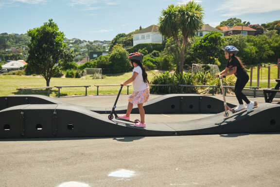 Two children wearing helmets ride their scooters on a wooden portable pump track in a school playground